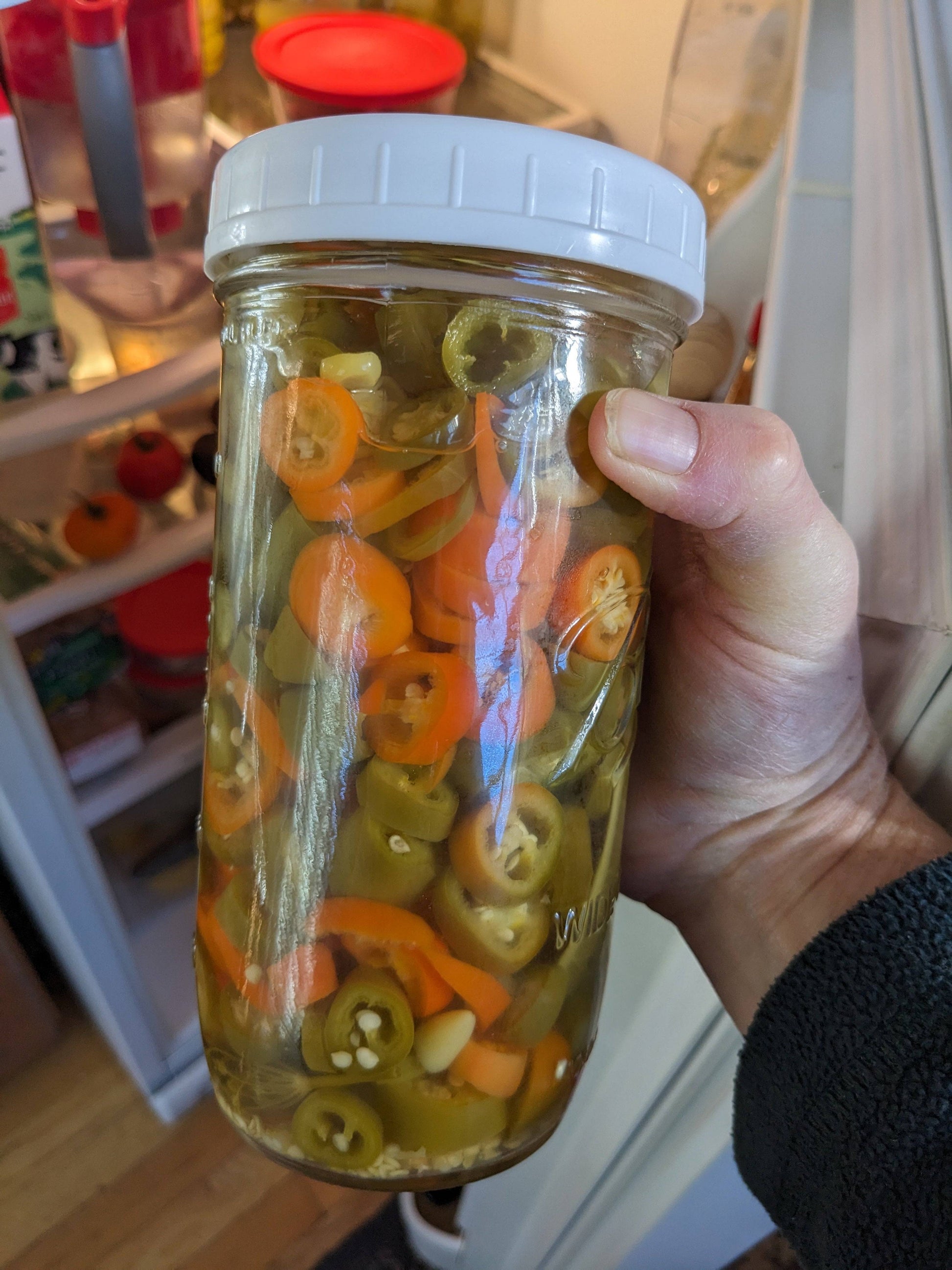 Jar of pickled NuMex Orange Spice jalapeños in a fridge. A spicy, colorful addition to homemade preserves and culinary creations.