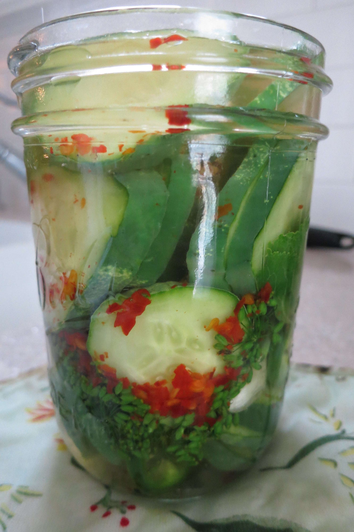 Glass jar filled with fresh cucumber slices, chopped herbs, and red pepper flakes, used for pickling and flavoring dishes