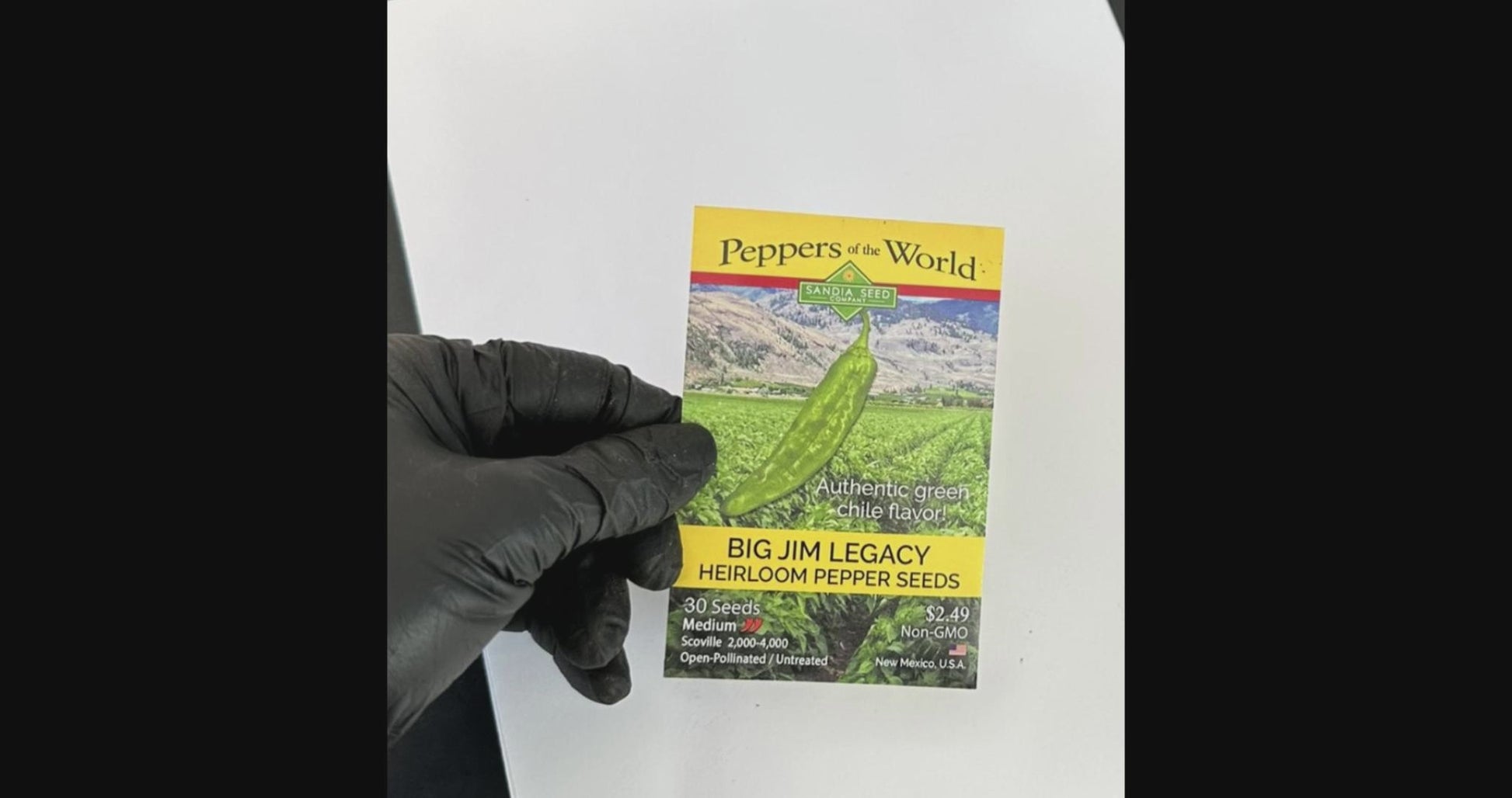Gloved hand holding Big Jim Legacy Pepper Seeds packet, emphasizing non-GMO heirloom seeds with medium heat and rich chile flavor.
