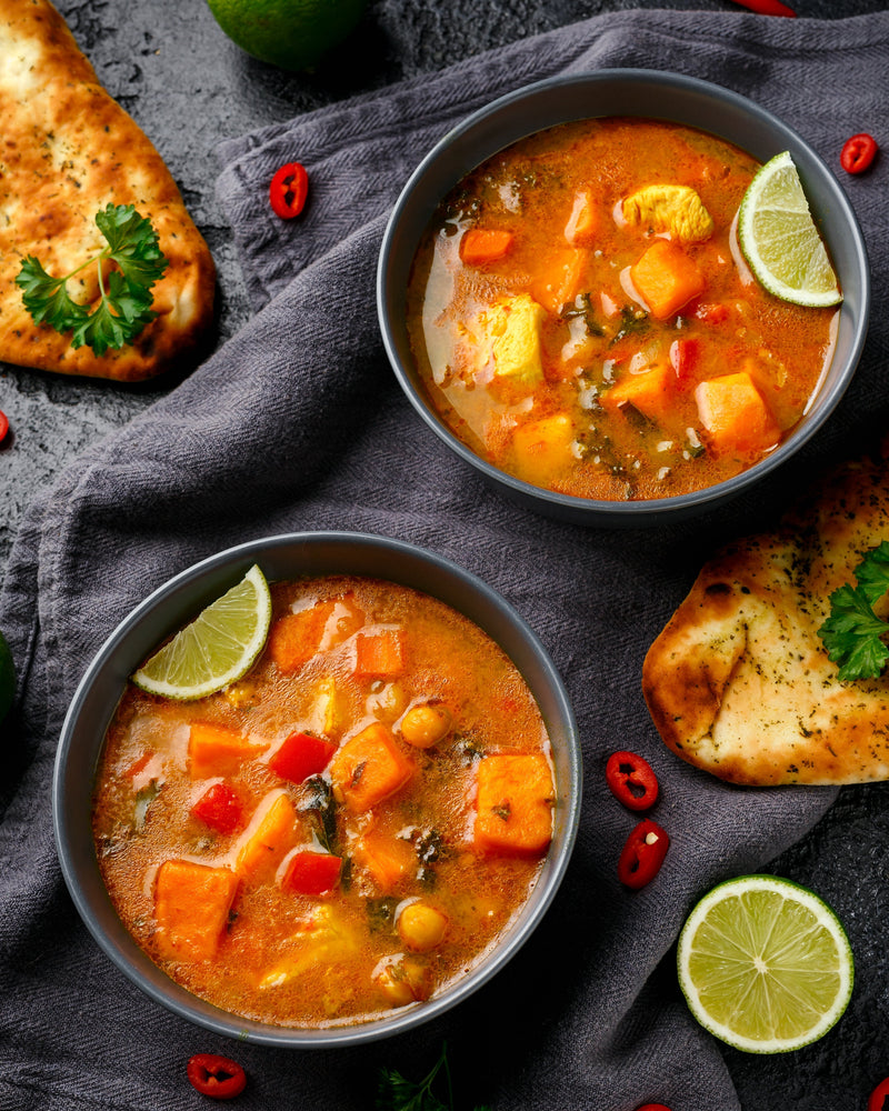 A bowl of hearty pumpkin chili soup with chunks of pumpkin and chickpeas, garnished with lime wedges, served alongside garlic bread on a dark textured surface.