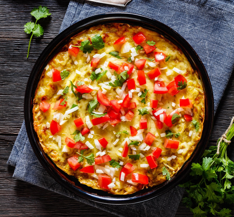 A baked green chile casserole topped with diced tomatoes, onions, and fresh cilantro, served in a black baking dish on a rustic wooden table.