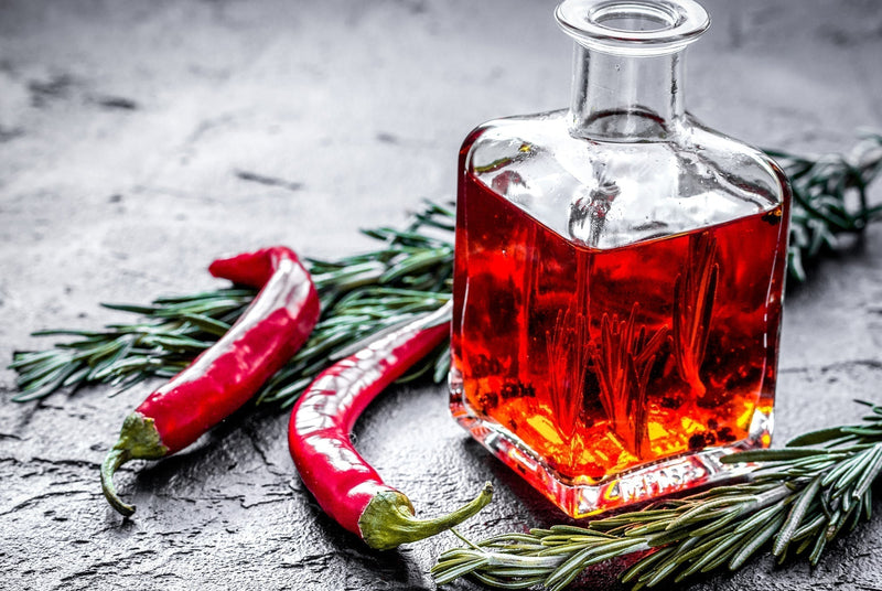 Chili Oil Recipe: A glass bottle of vibrant red chili oil surrounded by fresh red chili peppers and sprigs of rosemary, on a textured gray surface.