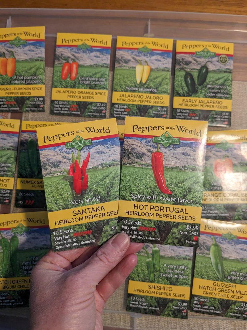 A close-up of seed packets for heirloom hot pepper varieties, including Jalapeño, Santaka, and Portugal, with vibrant peppers and scenic background.