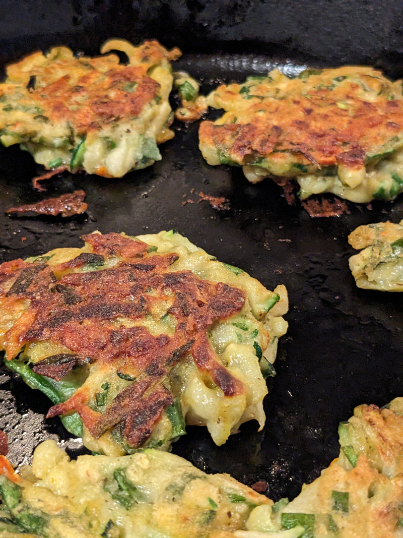A close-up of savory apple fritters cooking in a black skillet, showcasing crispy, golden-brown edges with visible green apple pieces and herbs.