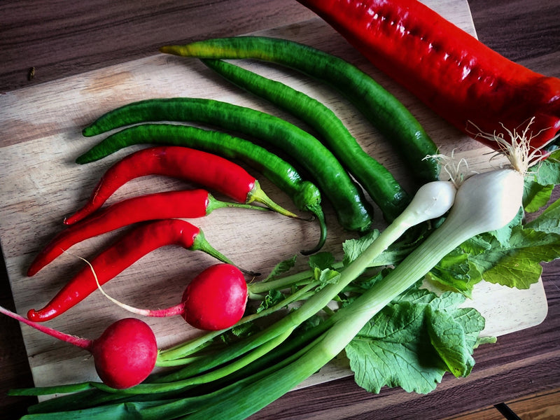 Gardening Questions? A colorful assortment of fresh garden vegetables including red radishes, green chili peppers, a red chili, and white spring onions on a wooden surface, ideal for gardening questions.