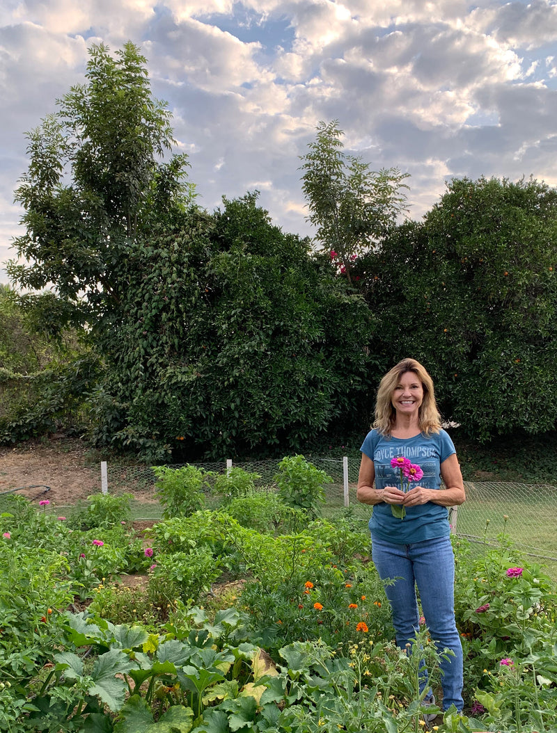 Should I Plant a Garden? Pictured: Sandia Seed owner, Patsy, smiling and holding flowers in a lush vegetable garden with vibrant flowers and vegetables, surrounded by green plants and trees under a partly cloudy sky.
