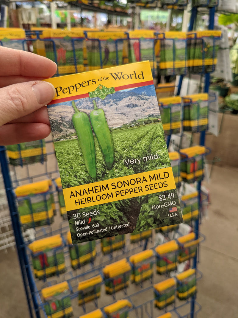 A close-up of a seed packet for Anaheim Sonora Mild heirloom pepper seeds, featuring vibrant green peppers and scenic farmland background, displayed on a store rack.