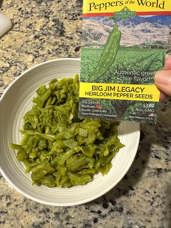 A hand holds a package of 'Peppers of the World' Big Jim Legacy Heirloom Pepper Seeds next to a white bowl filled with cooked green chili peppers on a granite countertop.