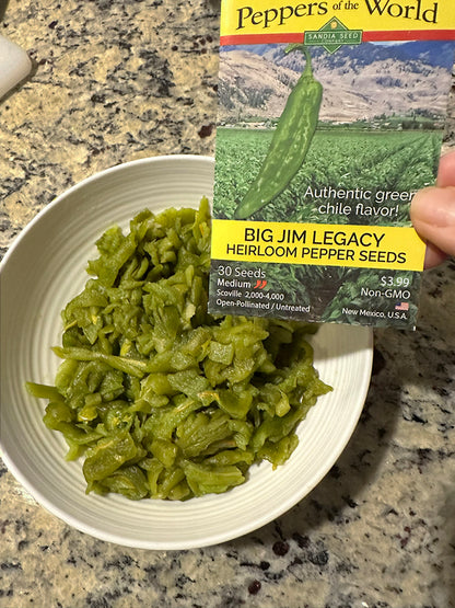 A hand holds a package of 'Peppers of the World' Big Jim Legacy Heirloom Pepper Seeds next to a white bowl filled with cooked green chili peppers on a granite countertop.