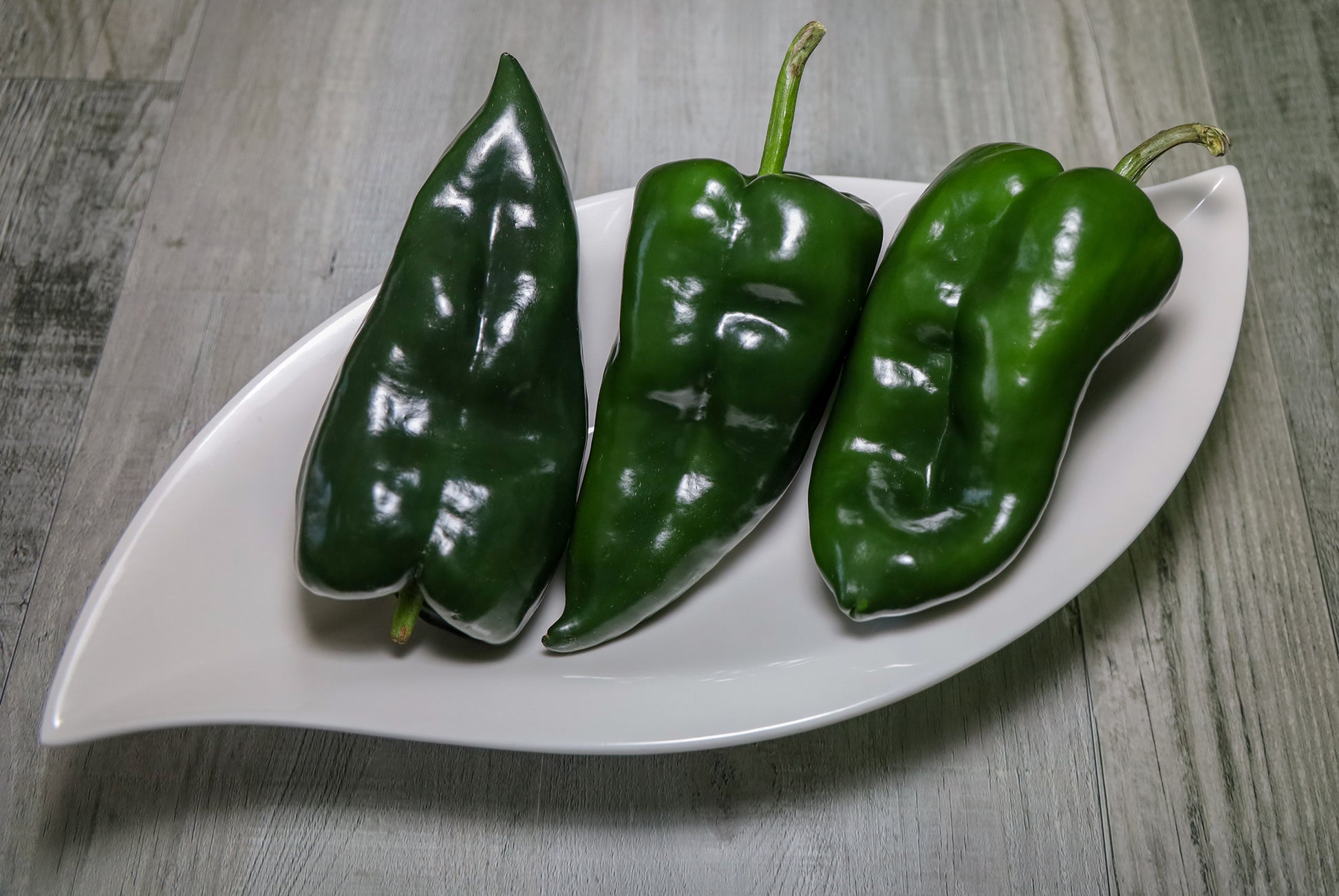 Three glossy green poblano ancho gigantea peppers arranged on a white leaf-shaped serving platter over a gray wood-textured surface.