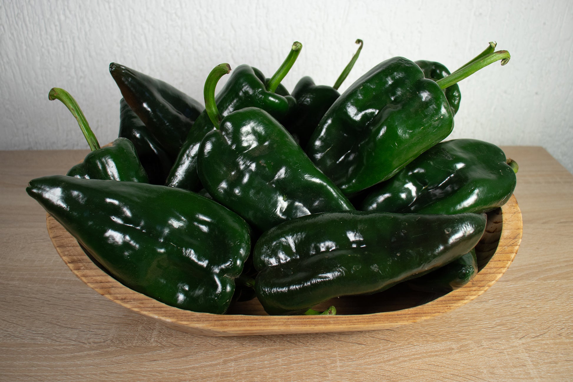 A pile of glossy dark green poblano ancho gigantea chili peppers in a light wood bowl on a wooden surface against a white wall.
