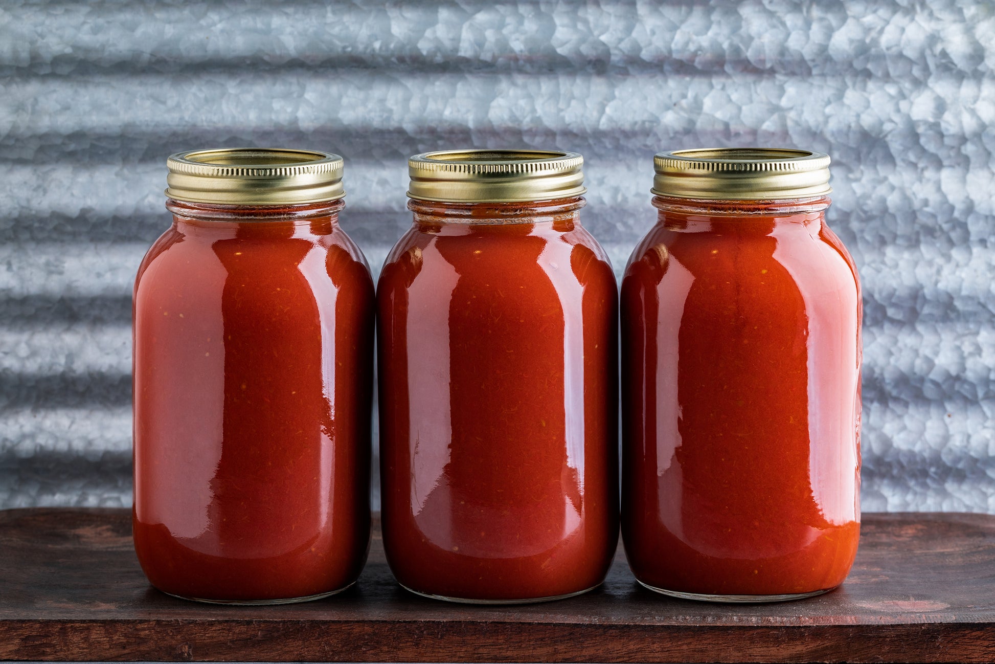 Three clear glass jars filled with red sauce, each with a gold metal screw lid, arranged side by side on a dark wooden surface against a textured gray corrugated metal background.