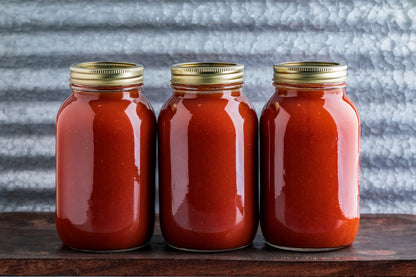 Three clear glass jars filled with red sauce, each with a gold metal screw lid, arranged side by side on a dark wooden surface against a textured gray corrugated metal background.