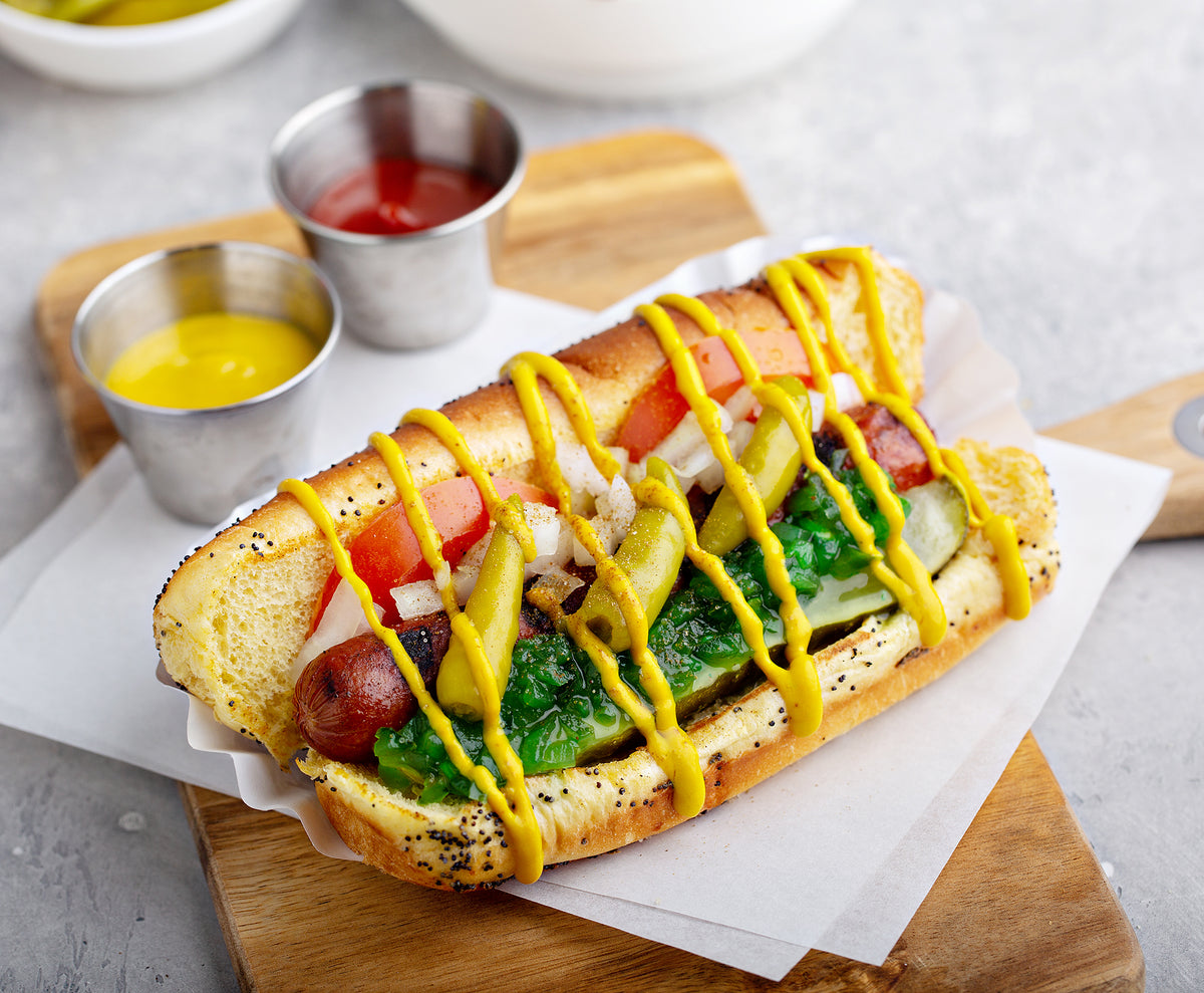 A gourmet hot dog in a poppy seed bun, topped with mustard, tomatoes, onions, pickles, and whole pickled sport peppers, served on a wooden board with small metal cups of mustard and ketchup on a light gray surface.