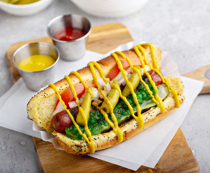 A gourmet hot dog in a poppy seed bun, topped with mustard, tomatoes, onions, pickles, and whole pickled sport peppers, served on a wooden board with small metal cups of mustard and ketchup on a light gray surface.