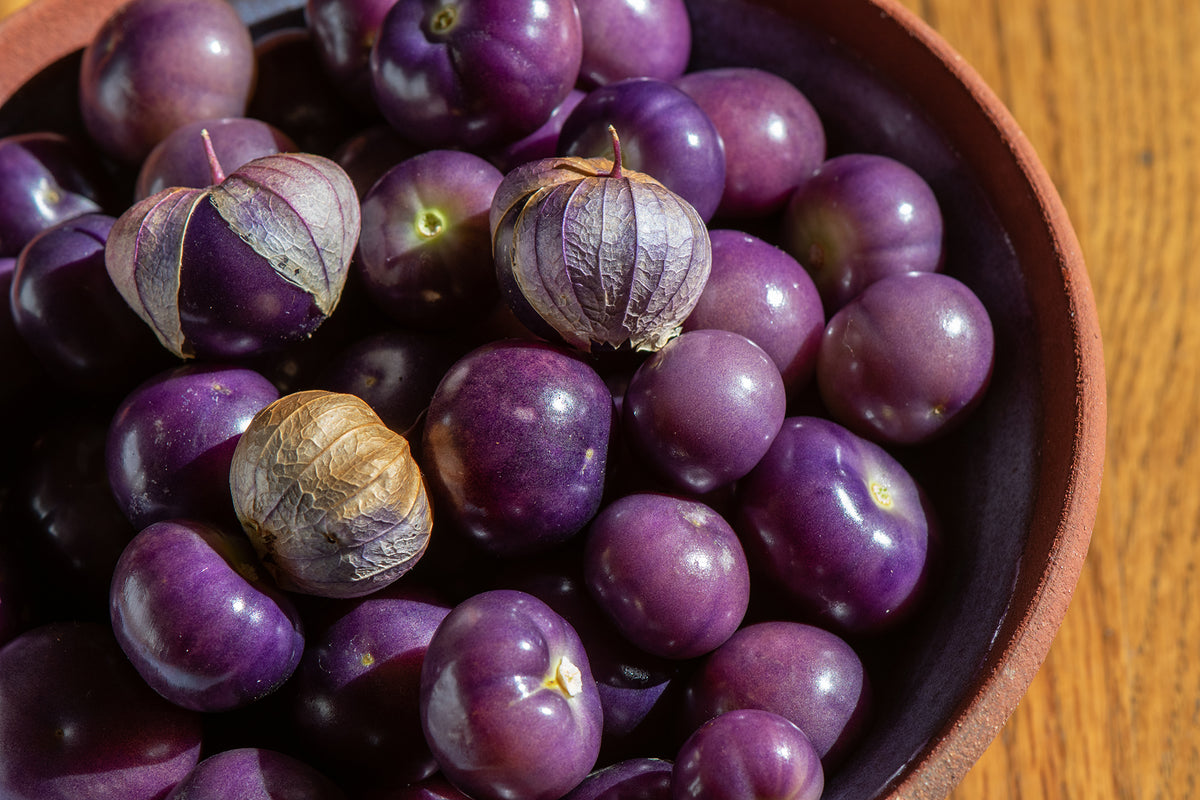 A close-up shot of purple goji berries, including one whole berry with its dried papery husk still attached, piled inside a dark bowl on a wooden surface.