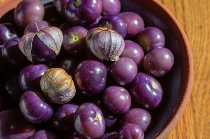 A close-up shot of purple goji berries, including one whole berry with its dried papery husk still attached, piled inside a dark bowl on a wooden surface.