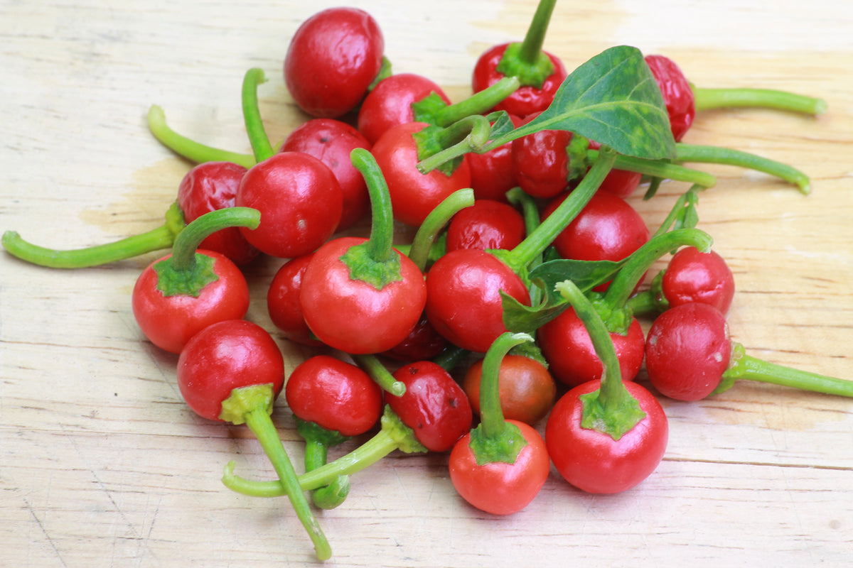 A pile of glossy sweet red cherry peppers with green stems and leaves, arranged on a light wood surface.